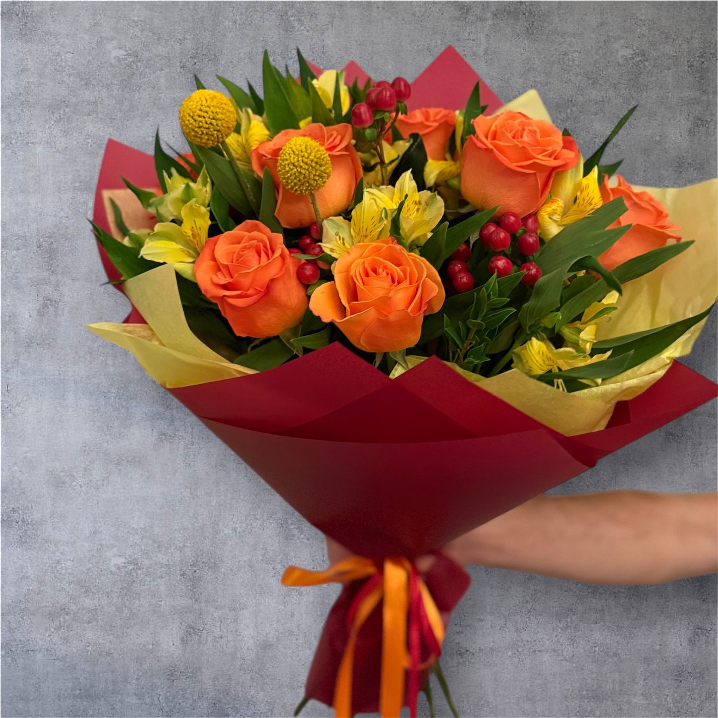 Bouquet of flowers with orange roses and yellow alstroemerias and craspedias with red hypericum wrapped in red paper, held against a gray background.