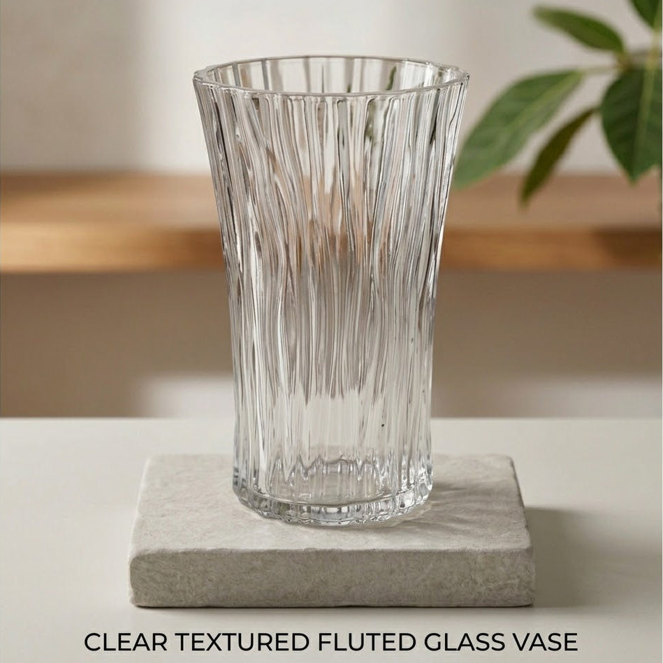 A clear textured fluted glass vase with a flared rim, displayed on a square stone coaster against a softly blurred indoor background with a wooden shelf and green leaves.
