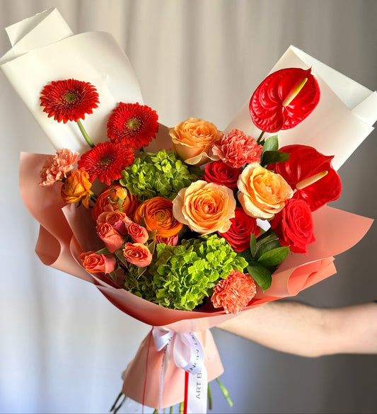 A vibrant hand-tied bouquet featuring bright red anthuriums, orange roses, and red gerbera daisies accented by lush green hydrangeas and peach carnations, wrapped in elegant light pink and white paper with a white ribbon.